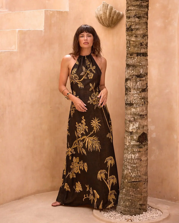 Woman in black halter maxi dress with gold floral embroidery, standing beside palm tree in warm-toned courtyard.