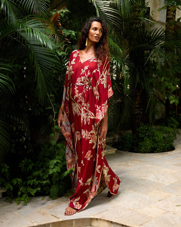 Woman in elegant red floral kaftan dress, walking barefoot on stone path amidst lush tropical greenery