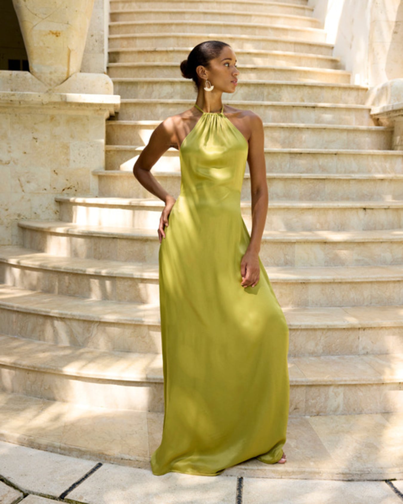 Woman in elegant olive green halter neck maxi dress, standing on stone steps, radiating confidence and grace.