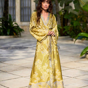 Woman in elegant yellow floral robe with birds, standing barefoot on stone patio amidst lush greenery