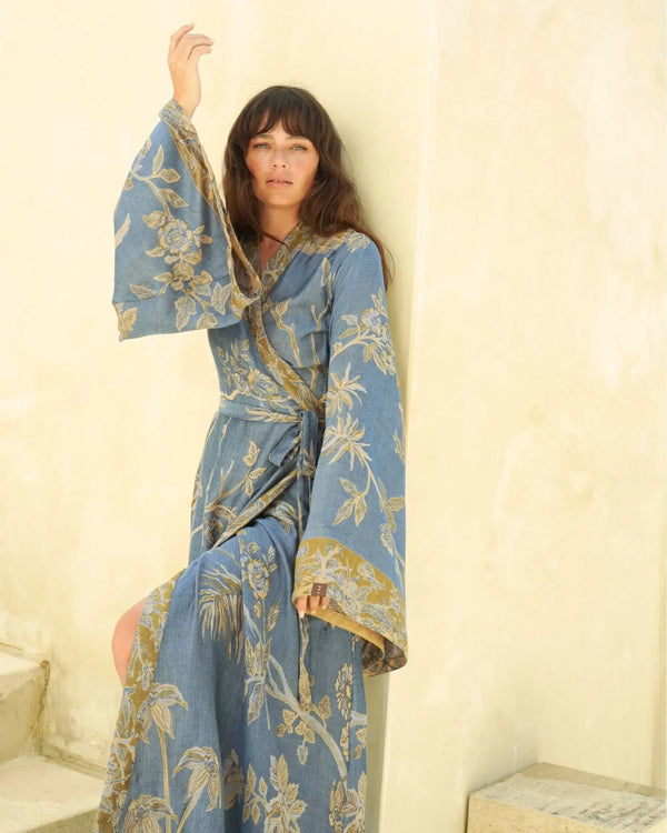 Woman in blue floral wrap dress with gold accents, standing against cream wall, boho style