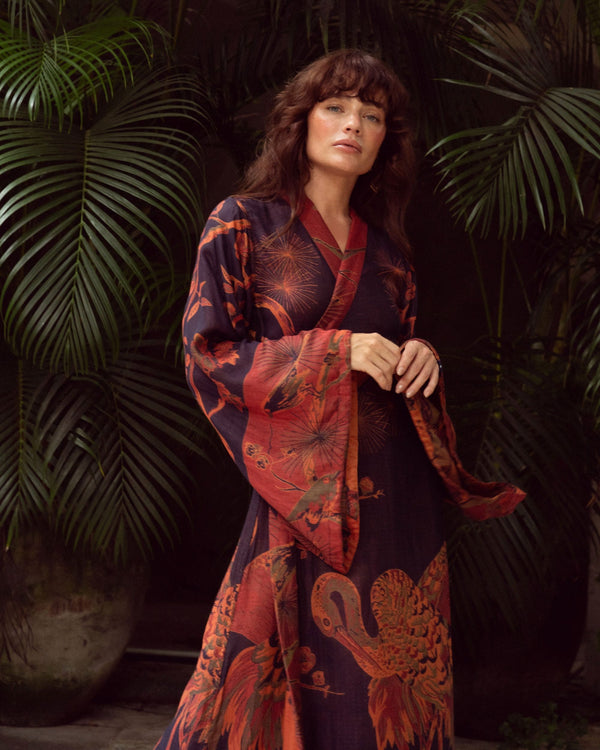 Woman in elegant, flowing robe with crane and floral print, standing amidst lush green palm leaves