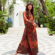 Woman in flowing orange floral robe, standing barefoot on patterned courtyard with lush greenery.