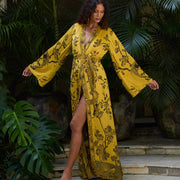 Woman in mustard yellow floral kimono dress with bird print, standing on stone steps amid lush tropical plants