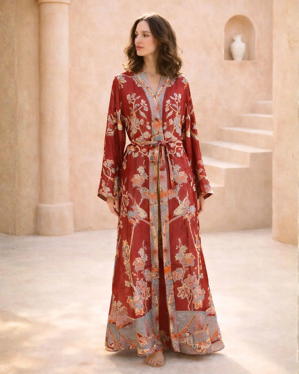 Woman in elegant red floral robe with long sleeves and tie waist, standing in sunlit courtyard