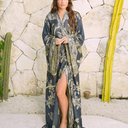 Woman in elegant floral robe with high slit, standing by stone wall and cacti, radiating confidence and grace.