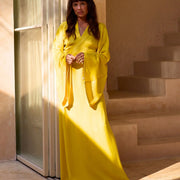 Woman in flowing yellow wrap dress, standing by sunlit doorway, elegant and radiant