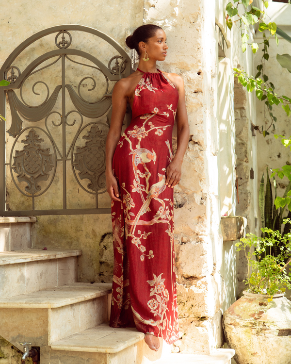 Woman in red floral halter maxi dress with bird print, standing by stone steps and iron gate